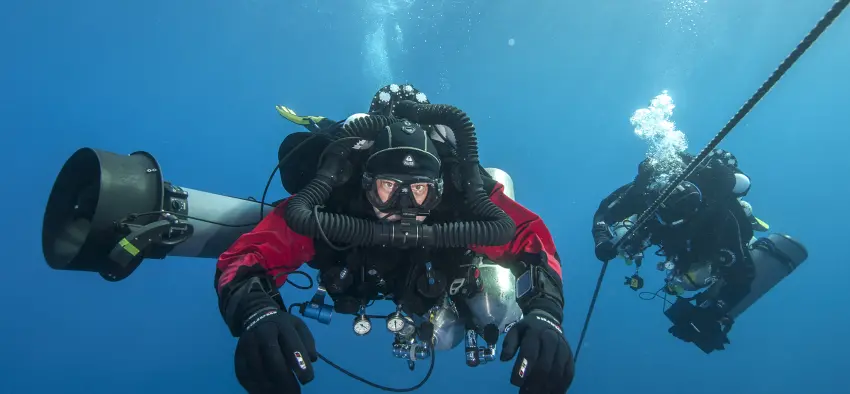 Technical diver in full gear with a Liberty closed-circuit rebreather, showing the oxygen cylinder, diluent cylinder, and breathing loop used for extended underwater dive time